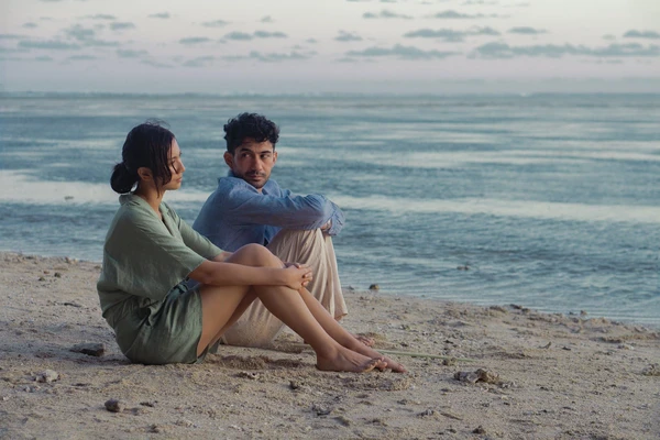 A man and a woman sit on a beach, sharing a moment in The Most Beautiful Girl in the World.